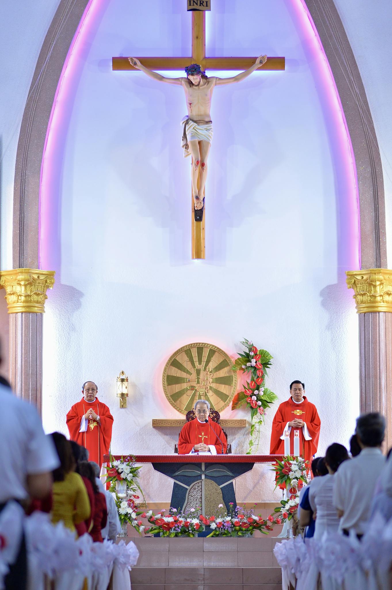 A solemn mass ceremony inside a decorated church with priests and a congregation, featuring a crucifix and vibrant lighting.